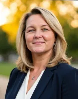 Professional woman with blonde hair wearing a navy blazer and white blouse outdoors with blurred greenery background