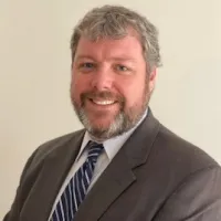 Smiling man with gray beard wearing a dark suit and blue striped tie against a plain background