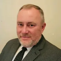 Middle-aged man with gray beard wearing a dark suit and polka dot tie smiling against light background