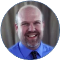 Smiling middle-aged man with bald head and gray beard wearing blue shirt and patterned tie.