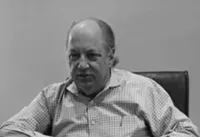 Older man in checkered shirt sitting in a chair against plain background with serious expression
