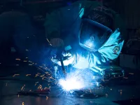 A welder in protective gear works on metal, creating bright sparks against a dark background.