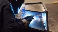 Welder wearing protective gear welding metal with sparks flying in a workshop environment.