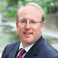 Portrait of a middle-aged man with glasses wearing a dark suit and red tie outdoors with blurred background