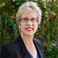 Smiling woman with short blonde hair, glasses, red lipstick, black blazer, and blue top, standing outdoors with greenery.