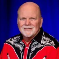 Smiling elderly man with white beard wearing red and black patterned shirt against blue background