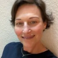 Close-up portrait of a smiling woman with short hair against a light textured wall, wearing a dark top.