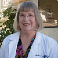 Smiling middle-aged woman wearing glasses and a white lab coat standing outdoors near greenery.