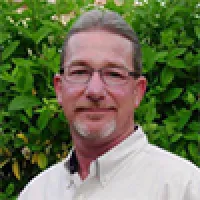 Middle-aged man with glasses and gray hair standing in front of green leafy plants wearing a white shirt