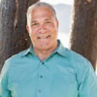 Smiling elderly man wearing a teal button-up shirt standing outdoors next to a tree.