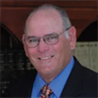 Smiling elderly man wearing glasses, a blue shirt, striped tie, and dark suit jacket in an indoor setting.
