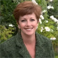 Smiling woman with short auburn hair wearing a green coat in a garden with white flowers in the background
