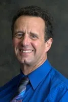 Middle-aged man with curly hair smiling, wearing a blue shirt and patterned tie against a dark background.