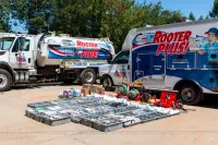 Plumbing work trucks and organized plumbing supplies laid out on pavement in an outdoor setting