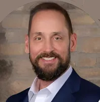 Smiling man with beard in a navy suit and white shirt standing against a stone wall background.