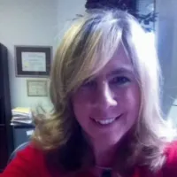 Smiling woman with blonde hair in red top sitting in office with framed certificates and bookshelf in background