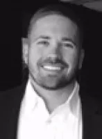 Smiling man with short hair and beard wearing a white shirt and dark jacket against black background
