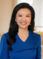Smiling woman with long black hair wearing a blue dress and pearl necklace in an indoor setting