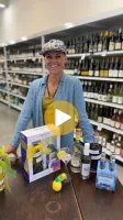 Smiling woman showcases cocktail kit and ingredients in a liquor store aisle filled with wine bottles.