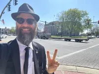 Bearded man in suit and hat making peace sign on city street with historic building and traffic lights in background