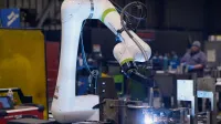 Industrial robotic arm welding metal parts in a factory setting with welding sparks visible.