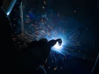 Welder wearing protective gear creates sparks while welding metal in a dark workshop environment.