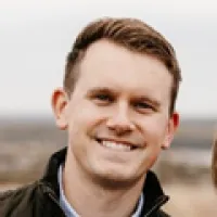 Smiling man with short light brown hair wearing a dark jacket outdoors with a blurred natural background
