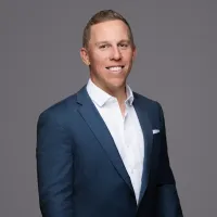Smiling man in a blue suit jacket and white shirt posing against a plain gray background.