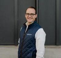 Smiling man wearing black glasses and a navy quilted vest with Nilsine Partners logo standing outdoors.