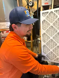 Estes Heating and Air Technician in orange shirt and blue cap installing a new air filter in a residential HVAC system.