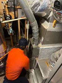 Estes Services Technician in orange shirt inspecting HVAC system components and ductwork in a home's utility room.