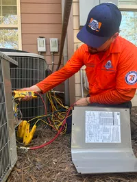 Estes Services HVAC technician in orange uniform using digital multimeter to service outdoor air conditioning unit with wiring and tools.