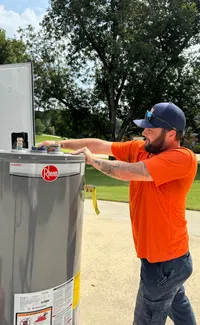 Estes Plumber in orange shirt installing or inspecting a Rheem water heater outdoors on a sunny day.