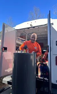 Estes Plumber in orange shirt working on a large water heater tank outside a service van on a clear sunny day.