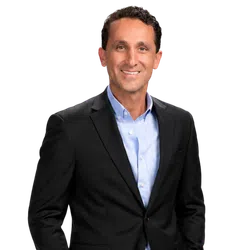Smiling man in black suit jacket and light blue shirt posing against a plain background.