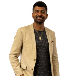 Smiling man wearing a beige blazer over a black patterned shirt with a gold pendant necklace