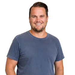 Smiling man with short hair wearing a blue t-shirt standing against a plain background.