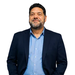 Smiling middle-aged man with beard wearing a blue shirt and navy blazer on black background.