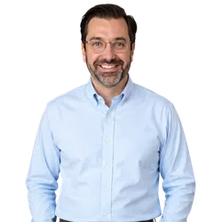 Smiling man with glasses and beard wearing a light blue button-up shirt against a white background