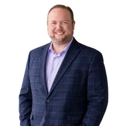 Smiling man wearing a blue plaid blazer and light purple shirt standing with hands in pockets.