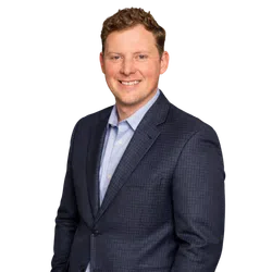 Smiling man with curly hair wearing a blue checkered blazer and light blue shirt against a white background