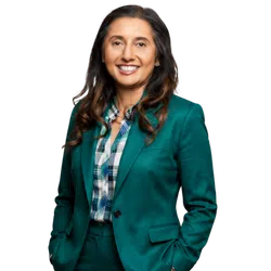 Smiling woman with long dark hair wearing a teal suit and plaid shirt posing confidently with hands in pockets.