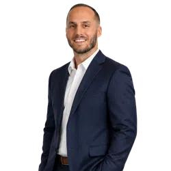 Smiling man in a navy blue suit and white shirt posing with one hand in pocket against a transparent background