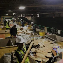 Workers in safety gear sorting materials on conveyor belts in an indoor recycling facility under bright lights