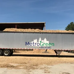 Metal trailer loaded with wood chips parked outside a recycling facility under blue sky.