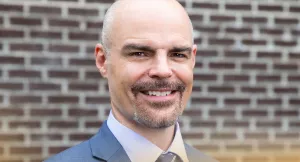 Smiling bald man with a goatee wearing a blue suit and tie standing in front of a brick wall outdoors