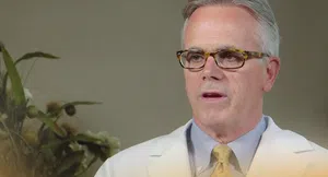 Middle-aged man in white coat and glasses speaking indoors with plants in the background.