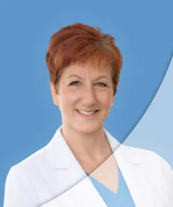 Smiling middle-aged female doctor with short red hair wearing a white coat and blue scrubs on blue background.
