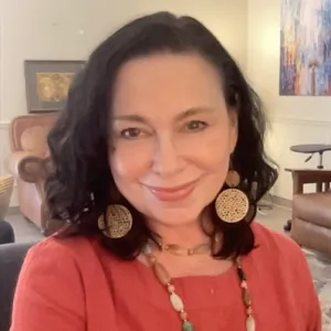 Smiling woman with dark hair wearing large round earrings and a beaded necklace in cozy living room