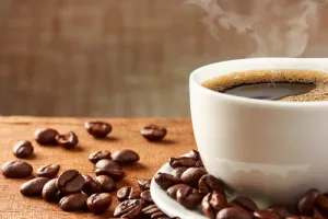 Steaming hot coffee in a white cup with roasted coffee beans scattered on a wooden surface.